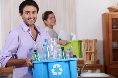 A man with dark hair, a beard, and a broad smile is holding a blue recycling bin filled with empty glass bottles, including clear, green, and brown varieties, in a domestic kitchen setting. The bin features a white recycling symbol printed on the side. Behind him, a woman with brown hair tied back, dressed in a light-colored top, is standing near a green storage box and appears to be engaging with the task at hand. The background includes wooden kitchen cabinets, a bamboo basket, and various household items, suggesting an organized and clean interior space. The lighting is bright and natural, highlighting the textures of the glass bottles and the smooth surfaces of the kitchen. The scene reflects an active effort in waste management, relevant to rubbish removal services such as those offered by House Clearance Farnborough, in a residential environment in Farnborough or nearby areas, with a focus on recycling and household cleanup activities.