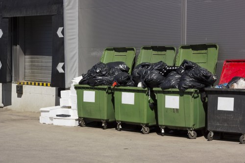 Three green wheelie bins with open lids are situated on a paved surface in front of a grey industrial building, filled with black rubbish bags. One red bin is partially visible nearby. The bins are placed on a concrete driveway or loading area, with some white bricks stacked to the left. The scene appears to be part of a waste collection or rubbish removal service in Farnborough, with the bins ready for collection or disposal. House Clearance Farnborough's name is associated with the image, highlighting their domestic property clearance services, and the environment is typical of a commercial or residential waste management setting in the local area. The lighting is natural, suggesting daytime, and the overall scene emphasizes waste removal readiness in a professional context.