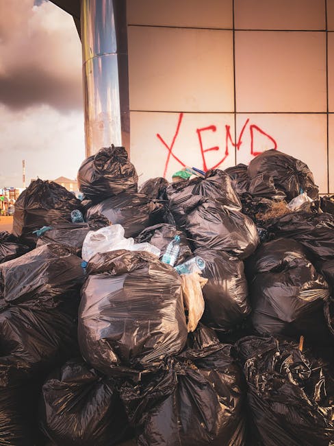 A waste collection worker wearing a bright orange safety vest with reflective stripes is seen operating a large red rubbish collection vehicle parked on the side of a street. The worker appears to be 