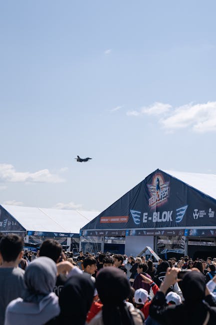 A military aircraft banked sharply in mid-air, leaving a white trail of contrail against a clear blue sky during the Farnborough Airshow. On the ground, three smaller aircraft with blue and white mark