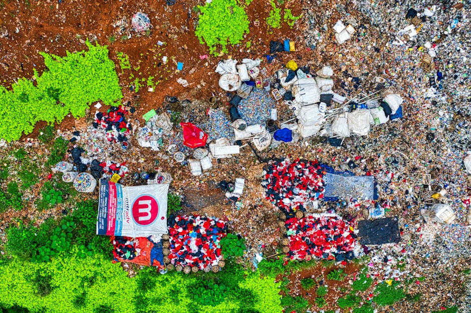 An aerial view of a large patch of land covered with a mixture of waste materials including plastic bottles, bags, and packaging, with some areas of exposed reddish-brown soil. There are scattered clu