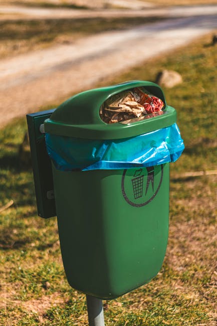 A large collection of mixed household waste and rubbish is piled on a paved pavement area in front of a parking space near a commercial building. The debris includes cardboard boxes, plastic bags, cru